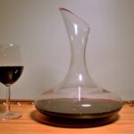 Dekanter mit breitem Boden, gefüllt mit Rotwein, neben einem Rotweinglas auf einem Holzbrett