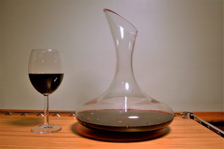Dekanter mit breitem Boden, gefüllt mit Rotwein, neben einem Rotweinglas auf einem Holzbrett