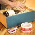 Weinflasche wird mit Holzwolle in einen Geschenkkarton gelegt, daneben Klebeband und Schere