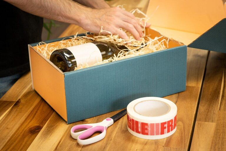 Weinflasche wird mit Holzwolle in einen Geschenkkarton gelegt, daneben Klebeband und Schere