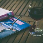 Glas Rotwein neben einer Lesebrille und einer aufgeschlagenen Zeitschrift auf einem Holztisch